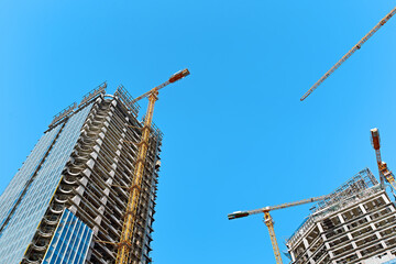 Construction of two skyscrapers with glasses with yellow tower cranes along the buildings in bright sunny weather against a clear blue sky, wide angle panoramic view from below