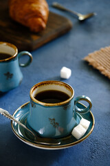 A crispy croissant with almond filling and hot black coffee on a wooden board on a blue concrete table.