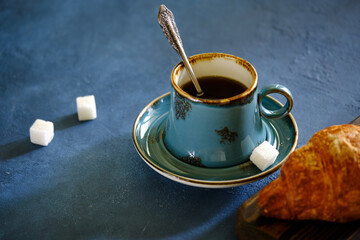 A crispy croissant with almond filling and hot black coffee on a wooden board on a blue concrete table.