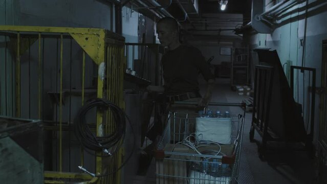 Medium long tracking of female survivor with shaved head pushing trolley with water bottles, looting in old warehouse with lights flickering