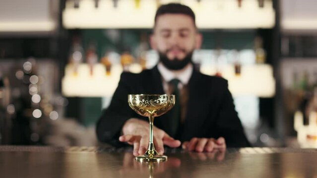 Blur background of competent bartender in stylish black suit putting on bar counter golden retro wine glass. Focus on antique shiny silverware used for alcoholic drink.