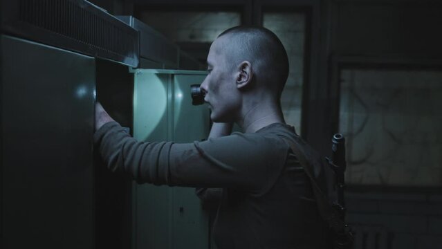 Medium tracking of bruised female soldier with shaved head holding flashlight, looting in abandoned dark locker room