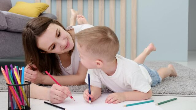 Millennial caring mother lying on floor with cute little child son, involved in creative activity at weekend time indoors. Happy young older sister or nanny having fun, drawing with small kid boy.