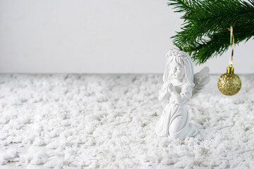 A white ceramic figurine of an angel on a light table. Spruce branch with a golden ball in the background. Snow on the table. Light background, space for text