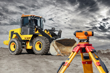 Bulldozer or loader moves the earth at the construction site against the sunset sky. Contrasting image of a modern loader or bulldozer. Geodetic equipment - a level for precise work.