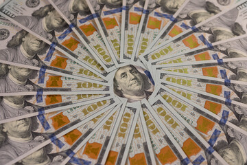 A close-up of many hundred-dollar bills lie in an even circle with Franklin's face in the center. Top view.