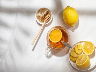 A mug of tea with lemons and a ginger root and little plate of honey with a wooden spoon on a table covered with a white tablecloth. Folk way of treating colds
