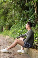 Vertical photo of athlete resting on the roadside. Young adult boy sitting resting after exercise with background full of greenery and nature.