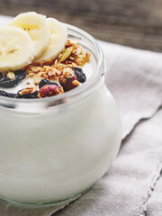 Natural homemade yogurt in a glass jar. Healthy food for breakfast with muesli. Jar with granola and banana slices on linen tablecloth on on wooden table.