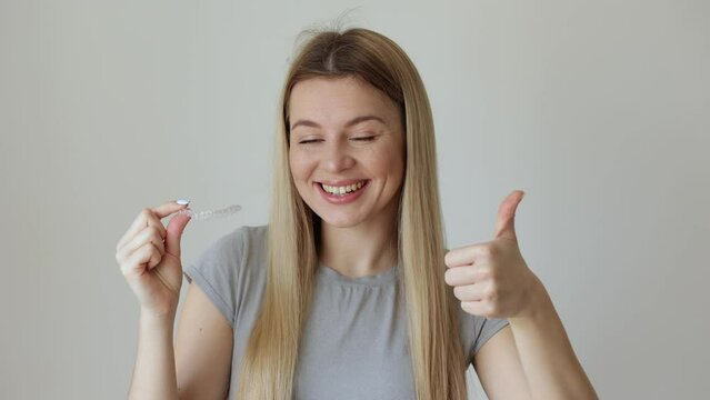 Happy young woman holding an invisible braces aligner and rising thumb up, recommending this new treatment. Dental healthcare concept.