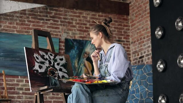 Young beautiful female artist painting still life with sunflower on canvas using oil paintings and art brush. Painter looking at the results of her work at art studio. Relaxation, leisure, hobby.