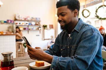 Black smiling man using mobile phone while having brunch in cafe