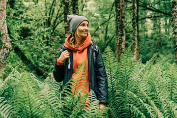 Young active woman hiker in a jacket and a hat with a backpack in a thicket of ferns in the forest