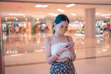 A young woman wearing a face mask inside a shopping mall checking her phone while walking around.