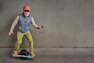 senior male hipster in helmet and goggles is riding one-wheeled electric skateboard in a grunge...