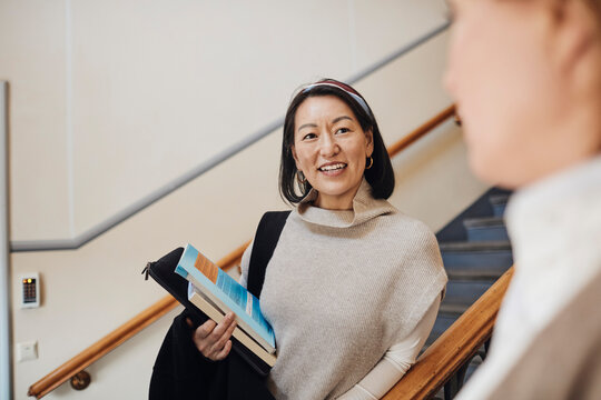 Smiling female teacher holding books while looking at colleague in university