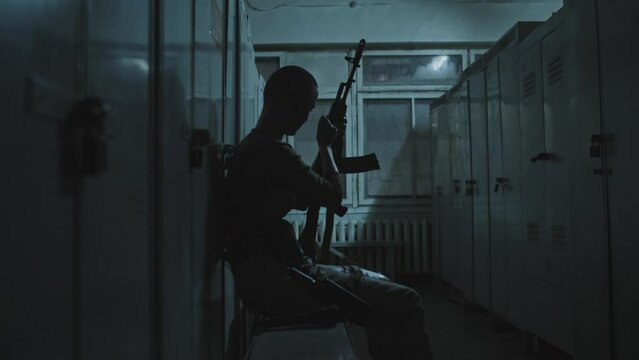 Medium long side view of female shooter with shaved head sitting in abandoned dark changing room, reloading shotgun