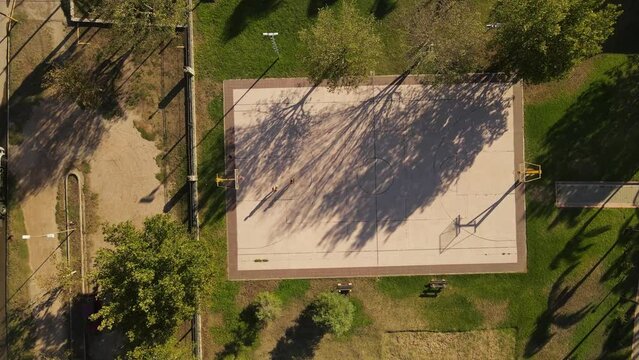 A dynamic aerial shot pivoting above a public basketball court while gradually descends to it. It's a public basketball court surrounded by trees and there are two persons playing basketball on it.