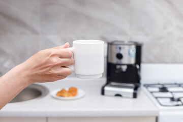 cup of coffee in hand background of a coffee machine and a croissant in the kitchen. coffee break