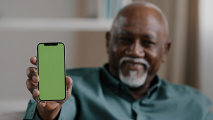 Old male African American biracial man at home sitting on couch businessman in office showing smartphone with empty green screen chromakey holding phone shows to camera advertisement gadget touchpad
