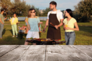 Empty wooden table and blurred view of friends cooking food on barbecue grill in park