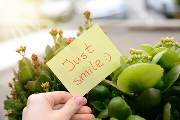 Woman holding paper note with handwritten text Just smile near beautiful plants against blurred...