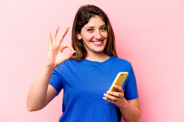 Young caucasian woman holding mobile phone isolated on pink background cheerful and confident showing ok gesture.