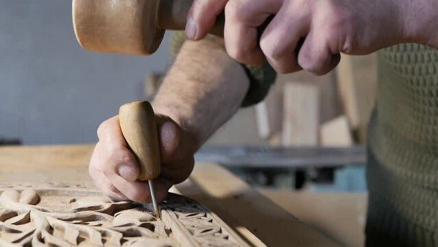 the male hands of a wood carving master holding a sharp awl with a wooden handle strikes with a wooden hammer and creates beautiful patterns on the board. Slow motion, close up. Hobby, craft