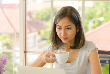 Smiling young asian woman wear casual clothing working with computer laptop and holding coffee mug in living room at home. WFH. Work at home concept.