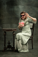 Comic portrait of young shocked girl in image of medieval royal person or princess in 3d eyewear with popcorn isolated on dark background.
