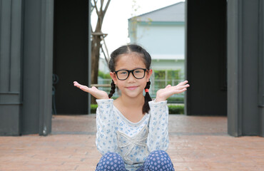 Asian little girl child wearing glasses with open hands and looking camera while sitting in pavilion at the garden outdoor.