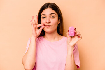 Young hispanic woman holding a keys car isolated on beige background with fingers on lips keeping a secret.