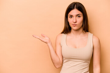 Obraz premium Young hispanic woman isolated on beige background showing a copy space on a palm and holding another hand on waist.