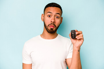 Young hispanic man holding car keys isolated on blue background shrugs shoulders and open eyes confused.