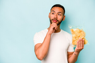 Young hispanic man holding a bag of chips isolated on blue background looking sideways with...