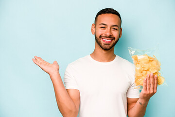 Young hispanic man holding a bag of chips isolated on blue background showing a copy space on a...