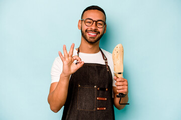 Young hispanic shoemaker man isolated on blue background cheerful and confident showing ok gesture.