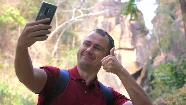 A smiling male traveler takes selfies on his phone against a canyon in a forest park. A happy man hiker records video or takes photos on camera with mountain canyons and river against forest backdrop.