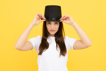 Teen girl in illusionist hat. Woman in magician top hat, black vintage cylinder cap on yellow background. Girl circus performer with headwear for magic tricks.