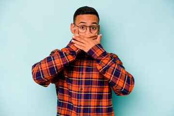 Young hispanic man isolated on blue background covering mouth with hands looking worried.