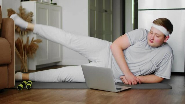 A chubby guy in sportswear, lying on a mat, performs exercises. The guy is watching a video tutorial on the laptop on how to properly perform the exercises. Sport