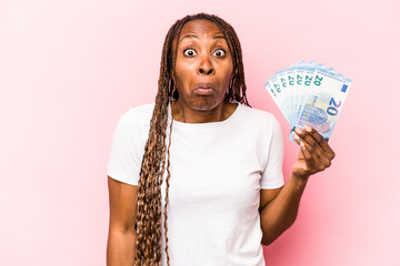 Young African American woman holding banknotes isolated on pink background shrugs shoulders and...