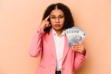 Young African American business woman holding banknotes isolated on beige background pointing temple with finger, thinking, focused on a task.