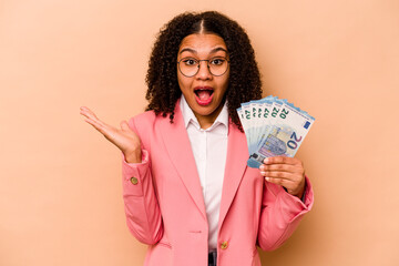 Young African American business woman holding banknotes isolated on beige background surprised and shocked.