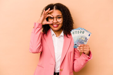 Young African American business woman holding banknotes isolated on beige background excited keeping ok gesture on eye.