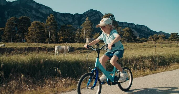 Cinematic footage of child boy ride bicycle on empty country road at sunset. Carefree children cycling on bike on village with mountain view. Happy childhood outside the town on holiday trip