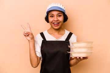 Young cook volunteer African American woman isolated on beige background joyful and carefree showing a peace symbol with fingers.