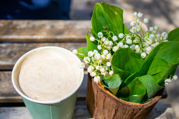 lily of the valley flowers in cute little pot on wooden table outside, in a public park.cappuccino drink and white flowers, people in the background. sunny spring,summer day. leisure concept