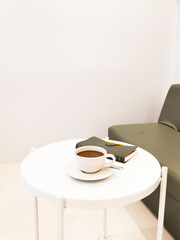 Coffee mug on white round table, there is a notebook and pen at the side and gray sofa on the right.