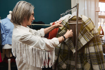 European mature woman, trendy fashion designer, tailor, seamstress standing in a garment storeroom, looking through the hangers with clothes ready for alteration.